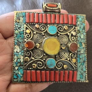 May include: A square pendant with a gold-colored frame, adorned with turquoise, red, and yellow stones. The design features intricate swirls and rectangular red accents, creating a detailed and colorful piece of jewelry.