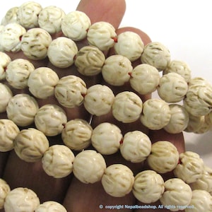 May include: A strand of off-white, spherical beads featuring a carved, textured floral pattern. The beads are strung together on a thin red cord, creating a continuous loop of round, patterned elements with a matte, organic appearance.