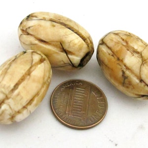 May include: Three oval-shaped beads with a cream-colored surface and dark brown veining, arranged around a copper-colored United States one-cent coin for scale. Each bead features carved vertical grooves along its length.