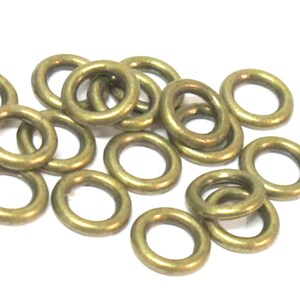 May include: A collection of small, brass-colored metal rings. The rings are circular with a hole in the center, and are scattered across a white surface. The rings have a slightly aged appearance.