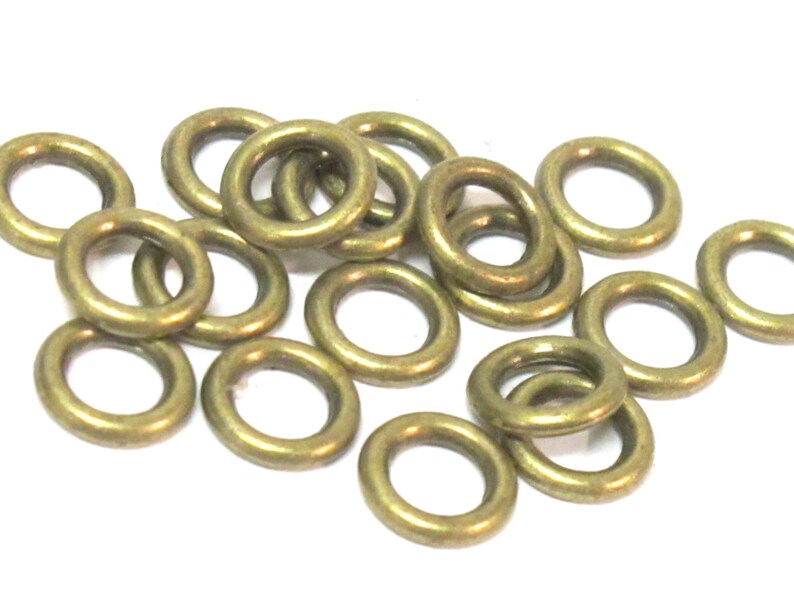 May include: A collection of small, brass-colored metal rings. The rings are circular with a hole in the center, and are scattered across a white surface. The rings have a slightly aged appearance.