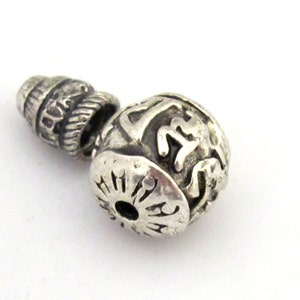 May include: A silver-colored bead with an intricate design. The bead has a cylindrical top with etched details, a central spherical section with a carved pattern, and a flared base with a circular opening. The bead is likely made of metal.