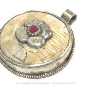 May include: Round pendant with a cream-colored, textured surface, silver-colored metal trim, and a flower design with a red center stone. The pendant has a loop for hanging.