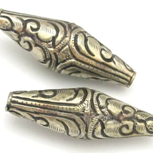 May include: Two silver-colored, elongated beads with intricate, swirling patterns. The beads have a cylindrical shape with tapered ends. The detailed designs are etched into the metal, creating a textured surface. The beads are displayed against a white background.