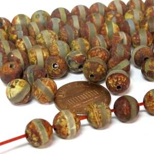 May include: A collection of round, rustic-looking beads with a mix of brown, yellow, and gray tones. Each bead has a lighter-colored band around the middle. A penny is included for size reference. The beads are strung on a red string.