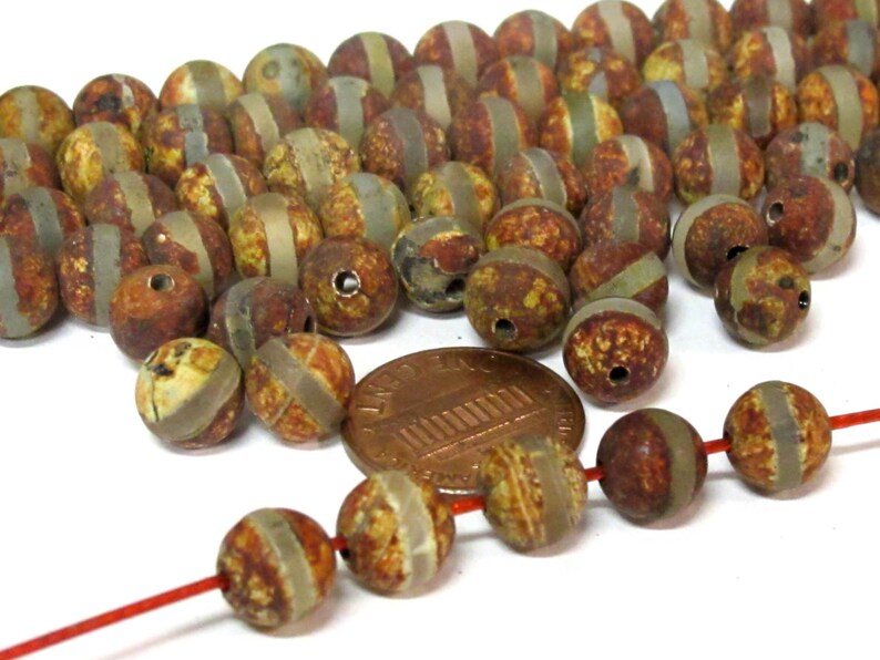May include: A collection of round, rustic-looking beads with a mix of brown, yellow, and gray tones. Each bead has a lighter-colored band around the middle. A penny is included for size reference. The beads are strung on a red string.