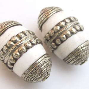 May include: Two barrel-shaped beads featuring smooth white centers contrasted by ornate silver-toned metal caps and a central band. The metal components display intricate etched cross-hatch patterns and a series of raised, rounded decorative studs.