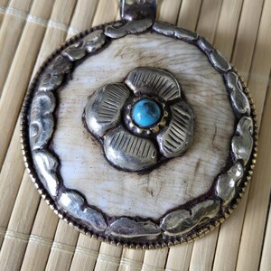 May include: A silver-toned pendant with a floral design and a turquoise stone center. The pendant has a circular shape with a decorative border and a bail for hanging. The background is a light-colored, textured surface.
