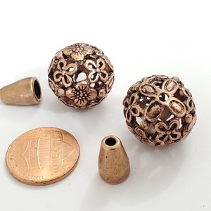 May include: Close-up of bronze-colored beads and components. Two spherical beads feature a floral design with openwork. Two cone-shaped beads and a penny are also visible. Ideal for jewelry making.
