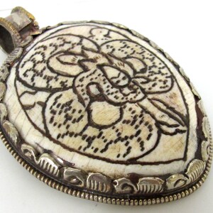 May include: Oval pendant with a cream-colored surface and a dark brown floral design. The pendant is framed with a silver-colored metal and has a silver-colored bail. The pendant is likely made of bone or a similar material.