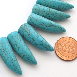 May include: Close-up of several turquoise-colored, elongated beads with brown veining. The beads are arranged in a row, with a penny for size comparison. The beads have a smooth, polished finish and a tapered shape.