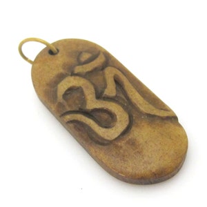 May include: A wooden pendant with a carved Om symbol. The pendant is a light brown color with a brass-colored loop at the top for hanging. The Om symbol is a deep brown color and is carved into the pendant.