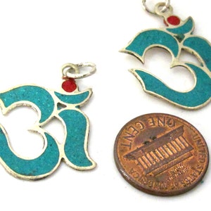 May include: Two silver-toned Om symbol pendants inlaid with turquoise-colored material and a small red dot at the top. Each pendant features a metal jump ring. A United States one-cent coin is placed next to the pendants for scale reference.