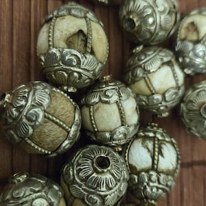 May include: Close-up of several decorative beads. Each bead features a cream-colored, spherical core encased in ornate, silver-toned metalwork. The metalwork has a floral design. The beads are arranged on a brown wooden surface.