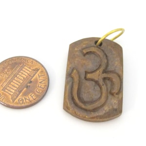 May include: A rectangular, brown pendant with the Om symbol carved into its surface. The pendant has a small, gold-colored loop at the top for hanging. A U.S. one-cent coin is next to the pendant for size comparison.