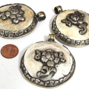 May include: Three round pendants with a cream-colored center and a silver-colored floral design. The pendants have a silver-colored border and a loop at the top for a chain. A penny is next to the pendants for size comparison.