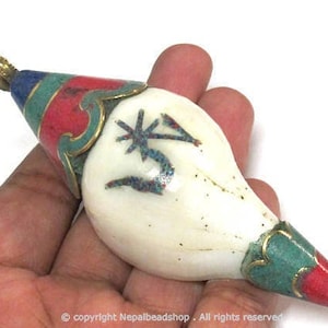 May include: A decorative pendant with a white, teardrop-shaped body. The pendant is adorned with red, green, and blue accents, and a gold-colored metal loop. The pendant has a unique design with a star and arrow symbol.