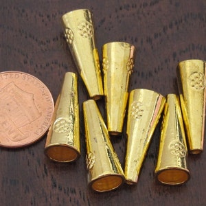 Op de afbeelding: Een verzameling goudkleurige, kegelvormige metalen kralen. Elke kraal heeft een decoratief bloemmotief aan de bovenkant. Een Amerikaanse munt is inbegrepen ter vergelijking. De kralen liggen op een donkerbruin oppervlak.