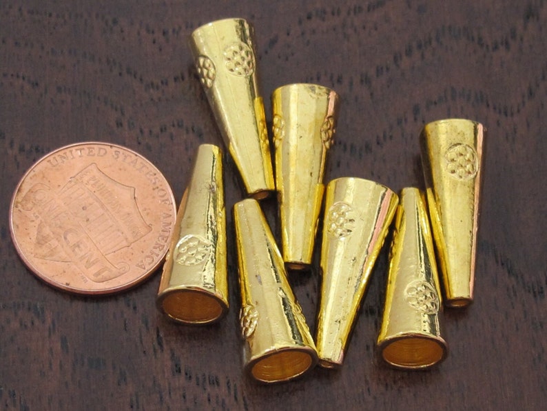 Op de afbeelding: Een verzameling goudkleurige, kegelvormige metalen kralen. Elke kraal heeft een decoratief bloemmotief aan de bovenkant. Een Amerikaanse munt is inbegrepen ter vergelijking. De kralen liggen op een donkerbruin oppervlak.