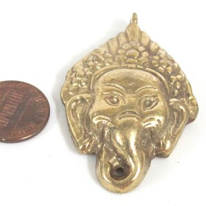 Puede incluir: Un colgante de lat&oacute;n con forma de cabeza de elefante, probablemente Ganesha, con detalles faciales y una corona texturizada. Una moneda sirve como referencia de tama&ntilde;o. El colgante tiene un lazo en la parte superior para colgar.