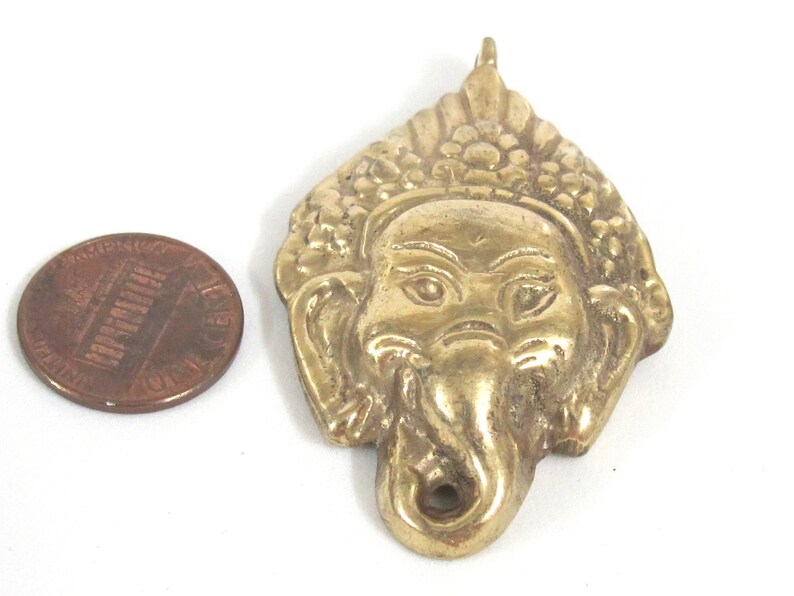 Puede incluir: Un colgante de lat&oacute;n con forma de cabeza de elefante, probablemente Ganesha, con detalles faciales y una corona texturizada. Una moneda sirve como referencia de tama&ntilde;o. El colgante tiene un lazo en la parte superior para colgar.
