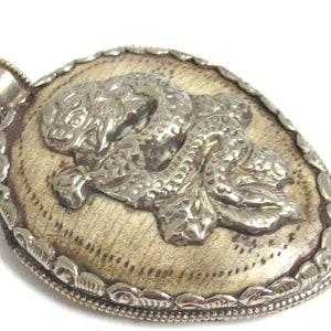 May include: Silver-toned pendant with a detailed relief of a mythical creature on a cream-colored background. The pendant has an oval shape with a decorative border and a loop for a chain.