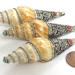 May include: Three decorative shell pendants with silver-colored metal accents. The shells are cream and brown, with turquoise and red accents. The pendants are cone-shaped and have intricate metalwork.