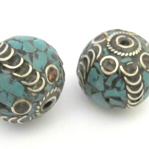 May include: Two round beads with a mosaic of turquoise-colored stones. The beads are accented with silver-colored metal, featuring curved and circular designs. The beads are likely for jewelry making.