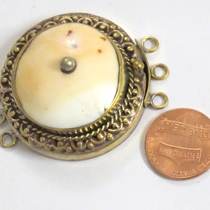 May include: A round, antique-style jewelry clasp with a cream-colored center and a decorative gold-toned metal frame. The clasp has three loops on one side and a small metal stud in the center. A penny is next to the clasp for size comparison.