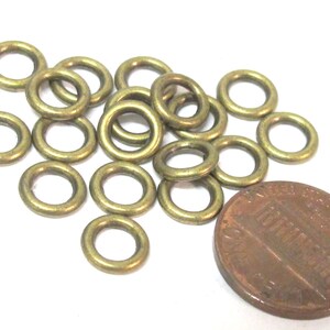 May include: A collection of small, brass-colored metal rings scattered on a white surface, next to a penny. The rings are circular with a hole in the center. The penny is brown and shows the profile of a person.