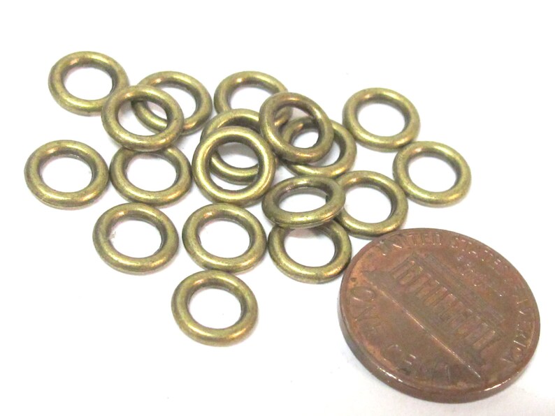 May include: A collection of small, brass-colored metal rings scattered on a white surface, next to a penny. The rings are circular with a hole in the center. The penny is brown and shows the profile of a person.