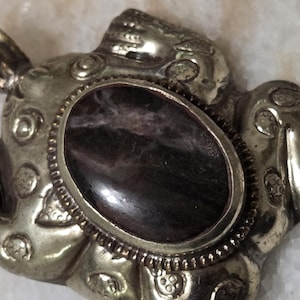 May include: A silver-toned pendant with an oval, dark gemstone center. The pendant has an ornate design with a loop for a chain. The gemstone has a swirling pattern of dark and light colors.