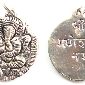 May include: A silver-colored pendant featuring the Hindu deity Ganesha. The front of the pendant shows a detailed relief of Ganesha, while the back has script. The pendant has a loop for a chain.