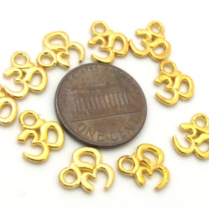 May include: A collection of small, gold-colored Om symbol charms. The charms are scattered around a penny, showcasing their size and detail. The Om symbol is a sacred sound and spiritual icon in Hinduism.