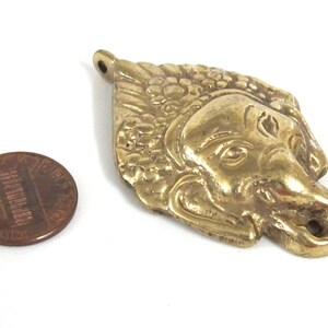 Puede incluir: Colgante de lat&oacute;n detallado con forma de rostro estilizado, posiblemente una figura mitol&oacute;gica. El colgante tiene detalles intrincados y un lazo para colgar. Una moneda est&aacute; al lado para la comparaci&oacute;n de tama&ntilde;o.