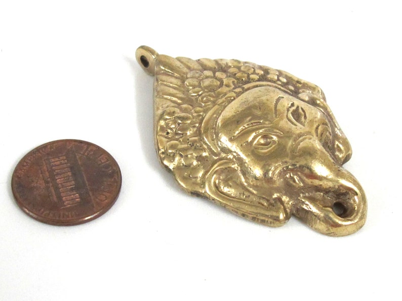 Puede incluir: Colgante de lat&oacute;n detallado con forma de rostro estilizado, posiblemente una figura mitol&oacute;gica. El colgante tiene detalles intrincados y un lazo para colgar. Una moneda est&aacute; al lado para la comparaci&oacute;n de tama&ntilde;o.