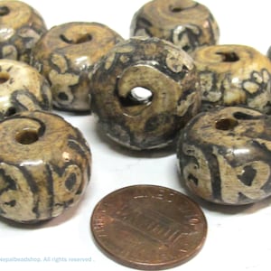May include: A collection of round, tan and dark brown patterned beads with a central hole, arranged near a copper-colored United States penny for scale. The beads feature a smooth, polished surface with intricate, dark, swirling etched designs.