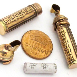 May include: A collection of antique gold-toned metal items, including two cylindrical pendants with engraved text, a small container, and a penny for size comparison. A small paper scroll is also present.