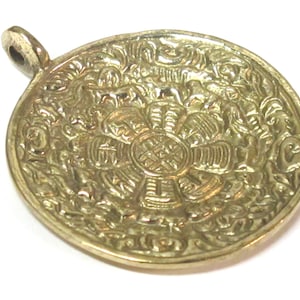 May include: A gold-colored, circular pendant with an intricate floral and geometric design. The pendant has a loop at the top for a chain. The detailed pattern covers the entire surface, creating a textured appearance.