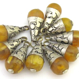 May include: A collection of amber-colored, teardrop-shaped pendants with silver-toned metal caps and loops. The pendants have a warm, translucent appearance, and the metal caps feature intricate decorative patterns. The pendants are arranged on a white surface.