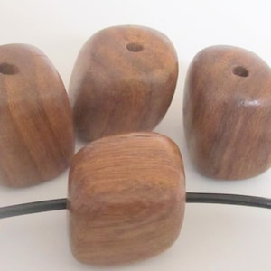 May include: Four smooth, brown wooden beads with a natural grain pattern. Each bead has a hole drilled through the center. One bead is strung on a black cord. The beads have a rounded, organic shape.