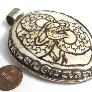 May include: Oval pendant with a cream-colored surface and a dark brown etched design. The pendant is framed with a silver-colored metal border. A penny is next to the pendant for size comparison.