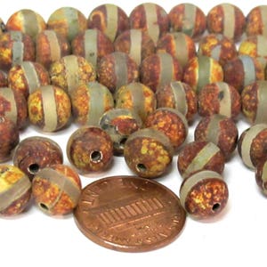 May include: A collection of round beads with a speckled, earthy color palette of brown, yellow, and green. Each bead has a band of light brown or tan encircling it. A penny is included for size reference.