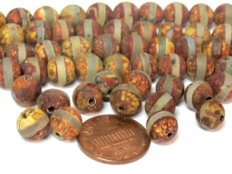 May include: A collection of round beads with a speckled, earthy color palette of brown, yellow, and green. Each bead has a band of light brown or tan encircling it. A penny is included for size reference.