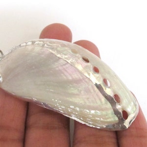 May include: A pearlescent abalone shell pendant with a silver-colored metal trim and bail. The shell has a natural iridescent sheen with visible holes along the side. The pendant is held in a hand against a white background.