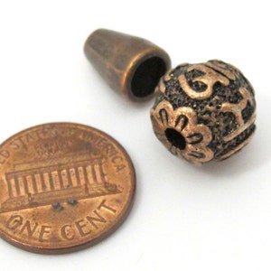 May include: Close-up of a copper-colored bead with an intricate floral design, next to a matching cylindrical clasp. A US one-cent coin is included for size reference. The bead has a detailed, textured surface, and the clasp is smooth.