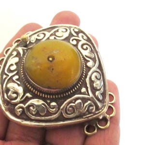 May include: Ornate silver-toned pendant with a large, round, yellow-orange stone at its center. The pendant has an intricate, swirling design around the stone and a decorative edge. A small gold-toned clasp is visible.