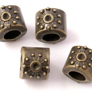 May include: Four antique bronze-colored metal beads. Each bead is cylindrical with a sunburst design and small dot accents. The beads have a hole running through the center, suitable for stringing.