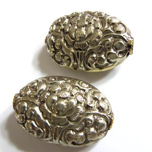 May include: Two oval-shaped, silver-colored metal objects with intricate floral designs. The objects have a raised, embossed pattern of flowers and leaves. The background is white, highlighting the details of the metalwork.