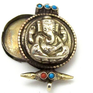 May include: A silver-colored pendant with a detailed relief of a deity, possibly Ganesha, within a circular frame. The pendant has a hinged compartment and is adorned with turquoise and coral-colored stones. The overall design suggests a religious or spiritual theme.